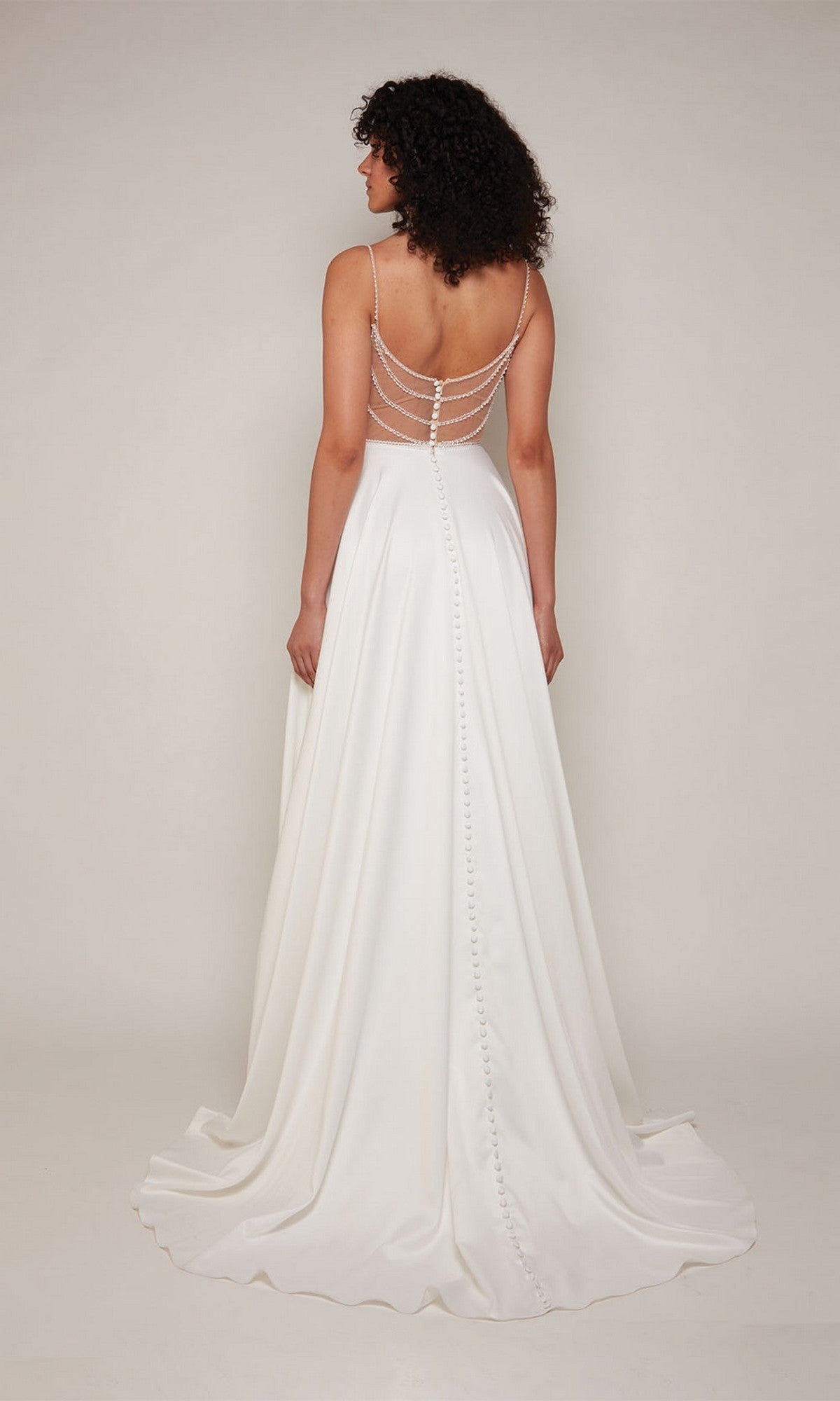 Wearing the Long White Dress: Alyce 7099, a woman stands facing away in a white satin formal gown with sheer beaded spaghetti straps, open back, and button details.